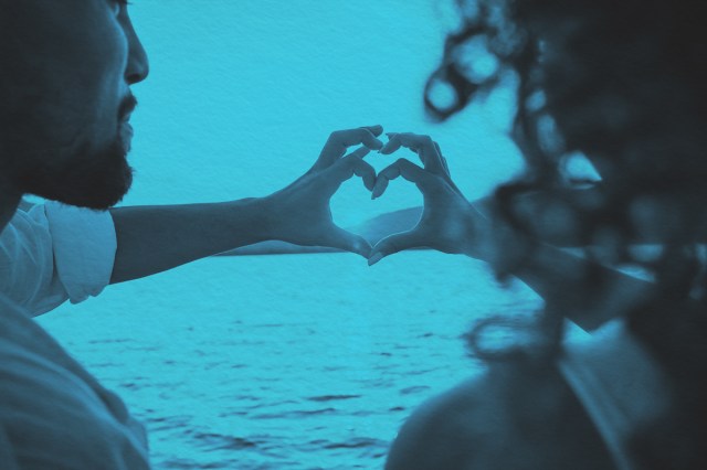 A couple doing heart shape with fingers looking at the sun setting behind the mountains